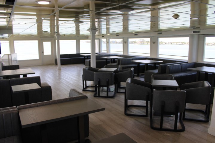Interior of an empty ferry lounge with tables, chairs, and large windows overlooking the water.