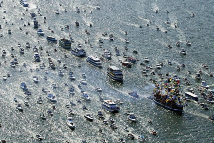 Numerous boats gather on a busy waterway, surrounding a large, colorful boat with flags.