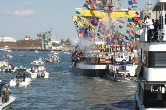 Decorated ship with colorful flags in parade, surrounded by smaller boats on waterway.
