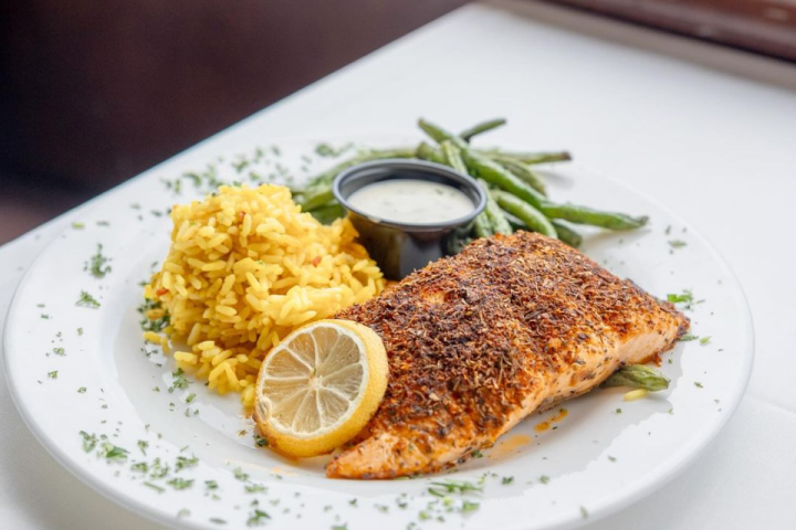 Seasoned salmon with lemon, yellow rice, green beans, and sauce on a white plate.
